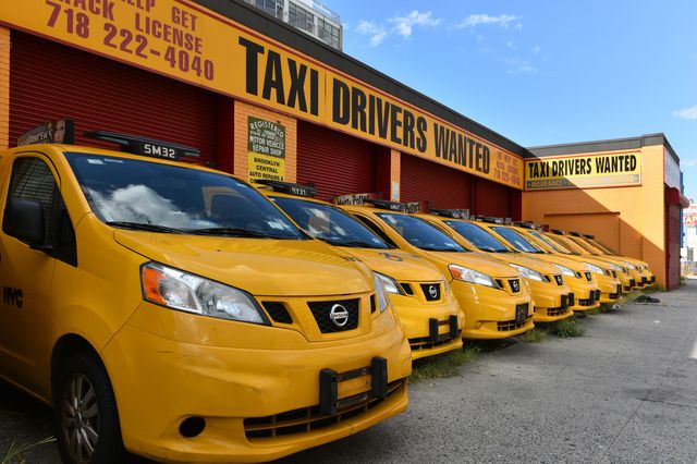 Dozens of taxies outside a taxi owner that has a store that says "Drivers wanted"